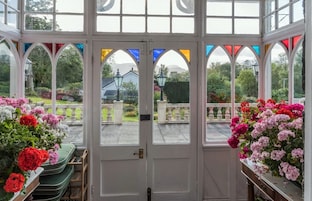 Un invernadero con puerta blanca, ventanas arqueadas y vistas a un jardín con flores y una casa al fondo.