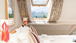 Un dormitorio con cama, almohadas, mesita de noche, lámpara, ventana con cortinas y vistas a montañas.