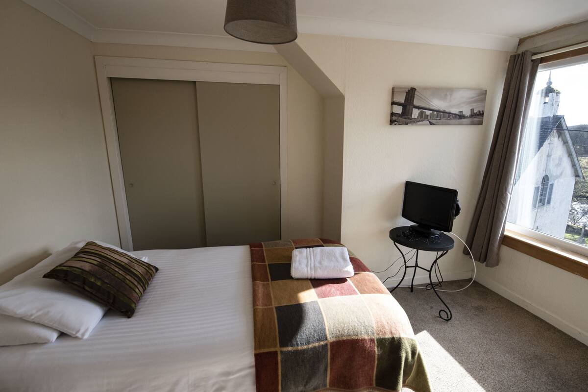 Een hotelkamer met een bed, een televisie op een klein tafeltje, en een raam met uitzicht op een gebouw.