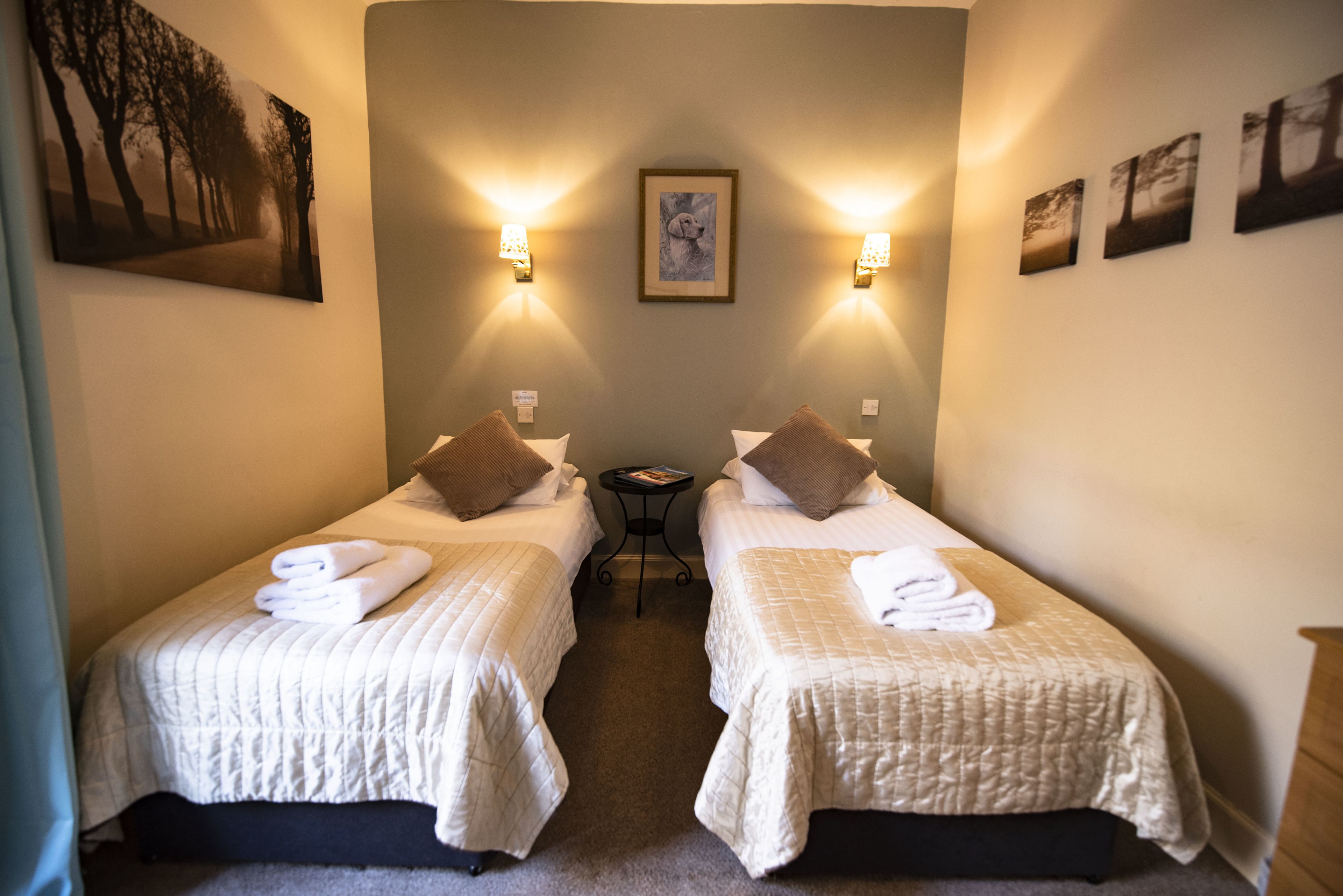 Habitación de hotel con dos camas, cada una con una almohada y una toalla enrollada. Hay una mesita entre las camas, y luces fijadas a la pared.