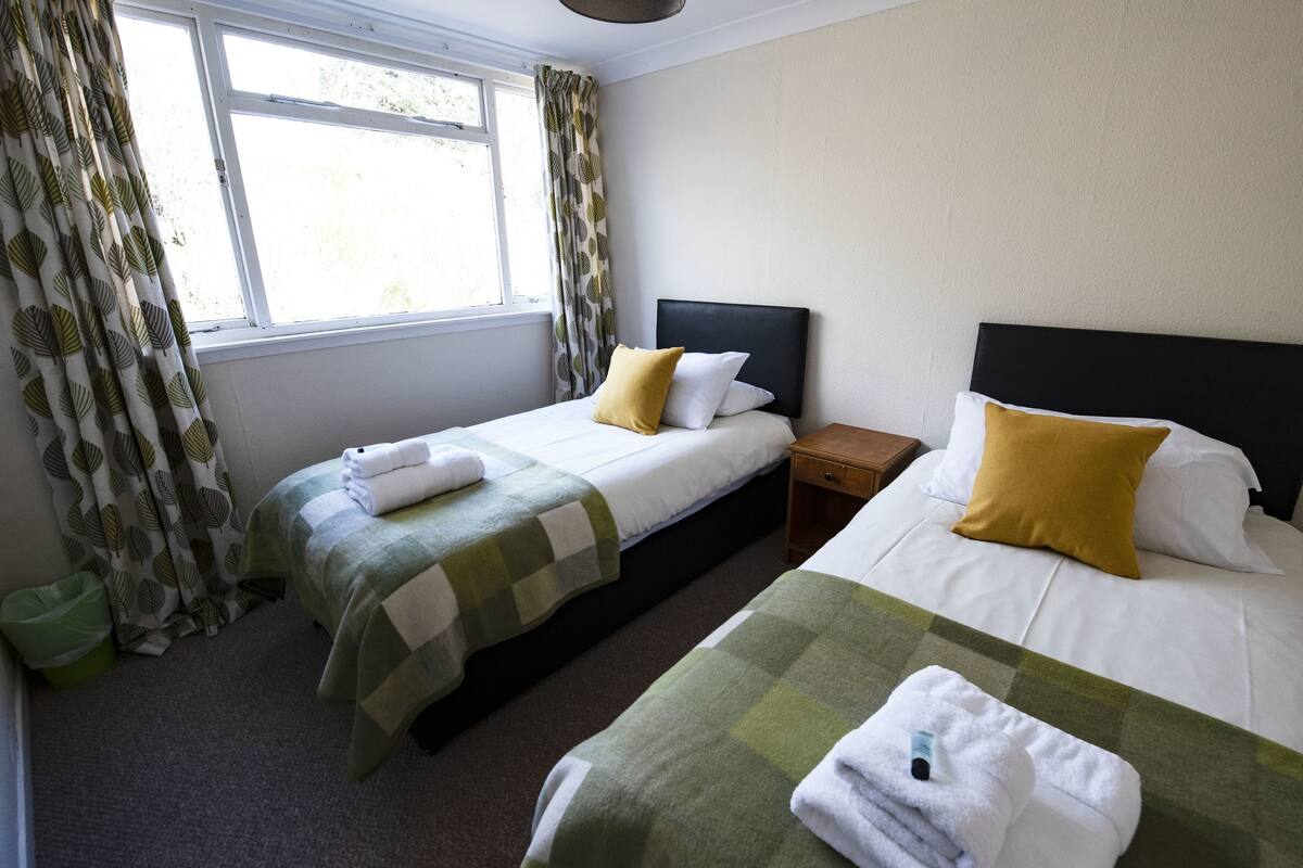 Een hotelkamer met twee bedden, een raam met gordijnen, een nachtkastje en een groene prullenbak.
