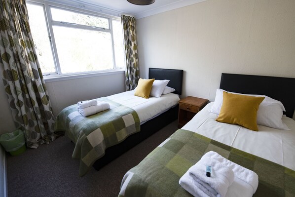 A hotel room with two beds, a window with curtains, a bedside table, and a green trash bin. - The Kirkmichael Hotel (Blairgowrie)