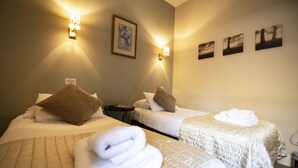 Habitación de hotel con dos camas, cada una con una almohada y una toalla enrollada. Hay luces fijadas a la pared y un cuadro enmarcado.