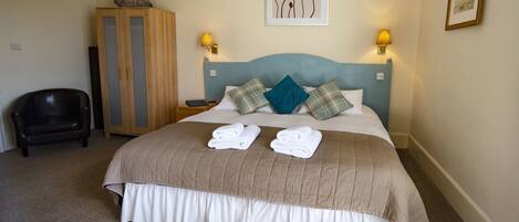 A neatly made bed with a headboard, two pillows, and two folded towels. There is a wooden wardrobe and a chair in the room.