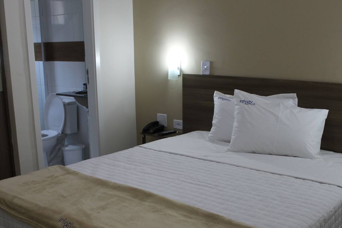 Une chambre d’hôtel avec un lit, une table de chevet, une lampe fixée au mur et une salle de bain visible à travers une porte ouverte.