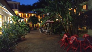 An outdoor patio area with tables and chairs, surrounded by lush greenery and a building with illuminated windows. - Parkview Inn (Moshi)