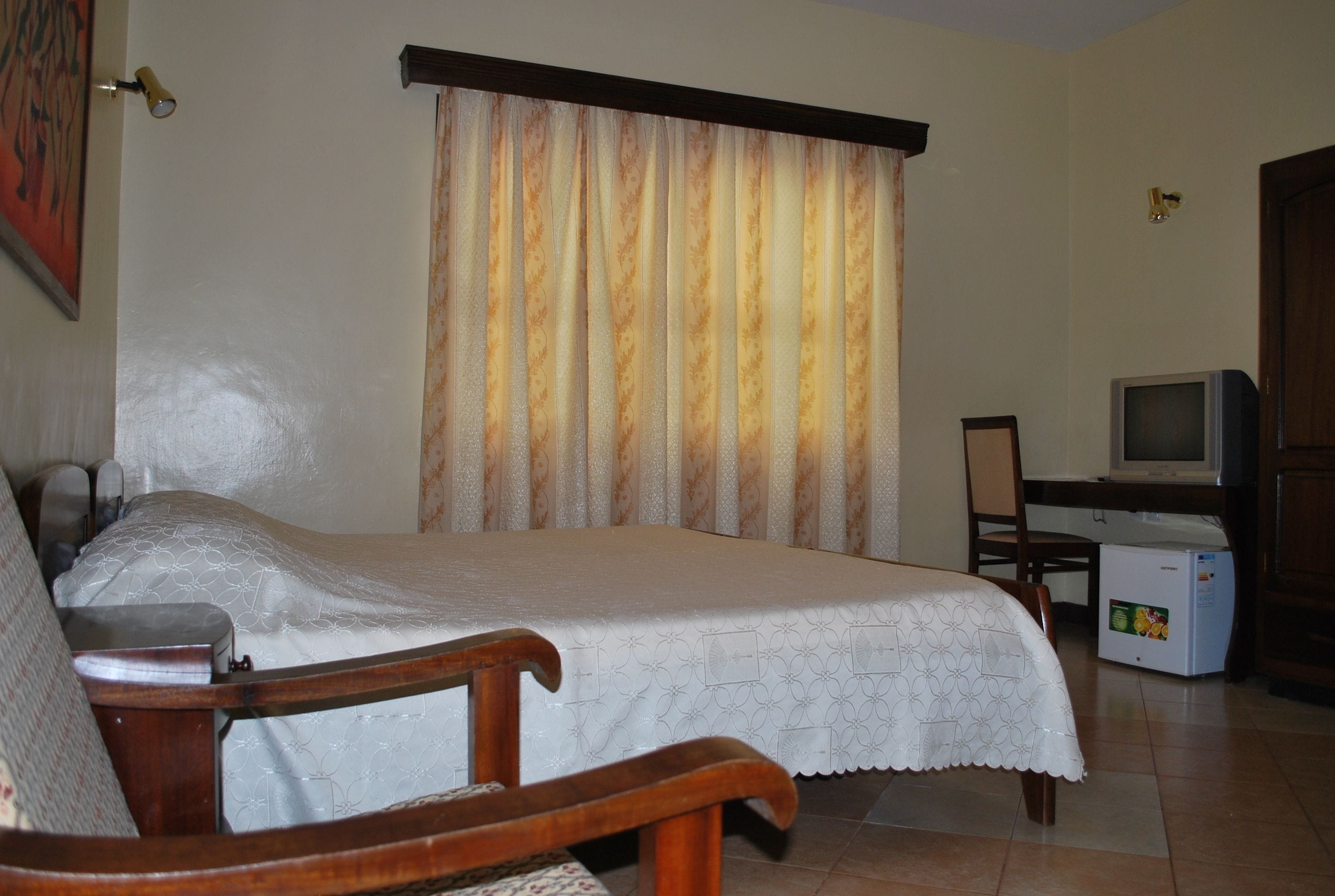 A bedroom with a bed, a wooden chair, a television, and a refrigerator.