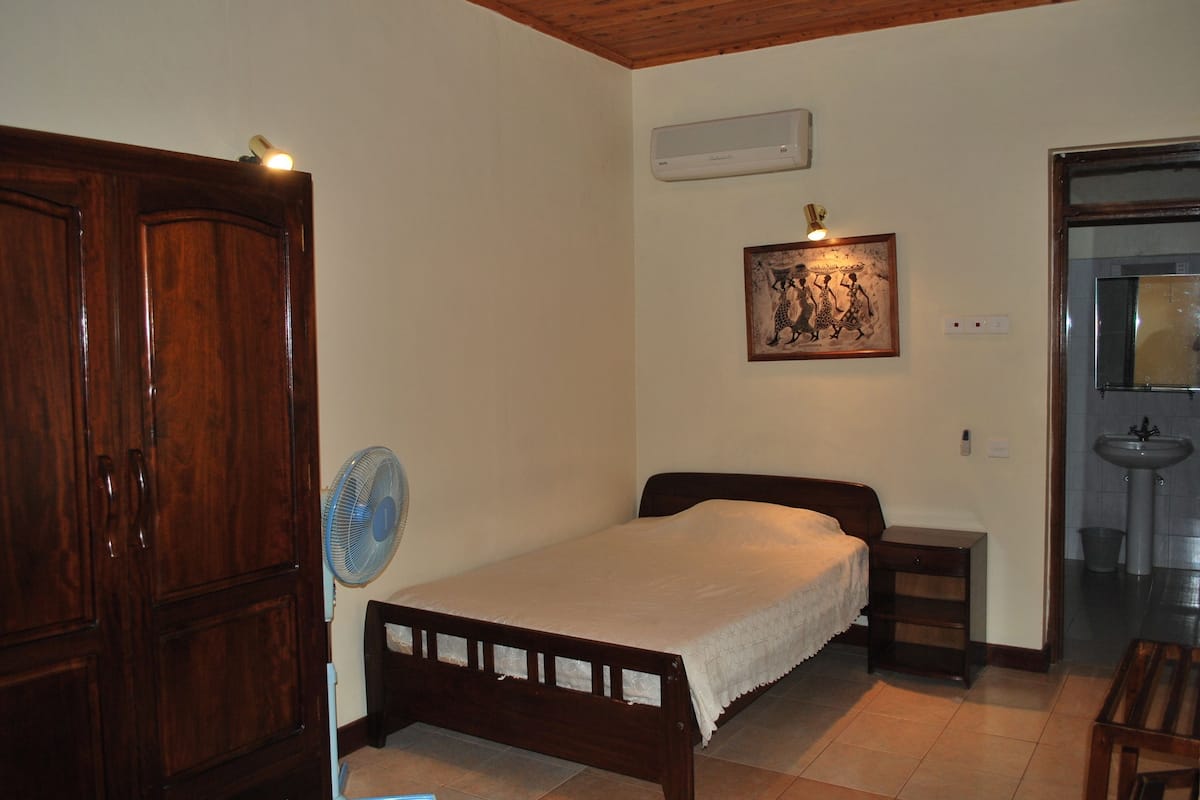 A bedroom with a wooden bed, a bedside table, a wall-mounted air conditioner, a wall-mounted fan, and a door leading to another room.
