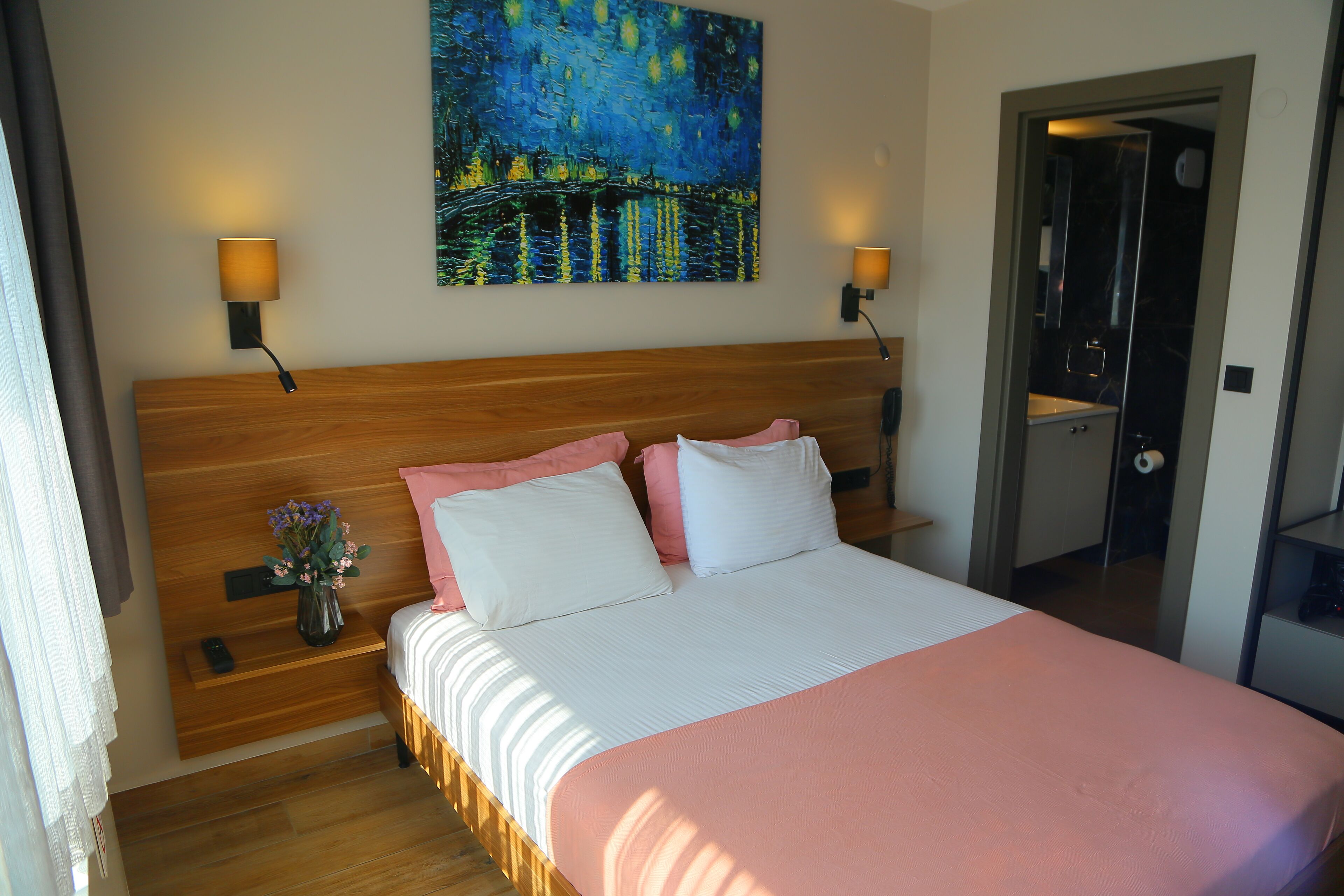 A bedroom with a wooden headboard, a bed with pink and white bedding, two wall-mounted lamps, a painting of a night scene, and a small vase with flowers on a nightstand.