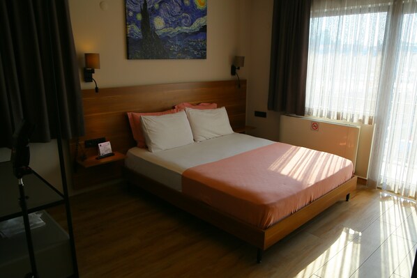 A hotel room with a wooden bed, a nightstand, a lamp, a painting on the wall, and a window with curtains.