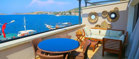 A balcony with a round table and wicker chairs, overlooking a harbor with boats and a distant coastline.