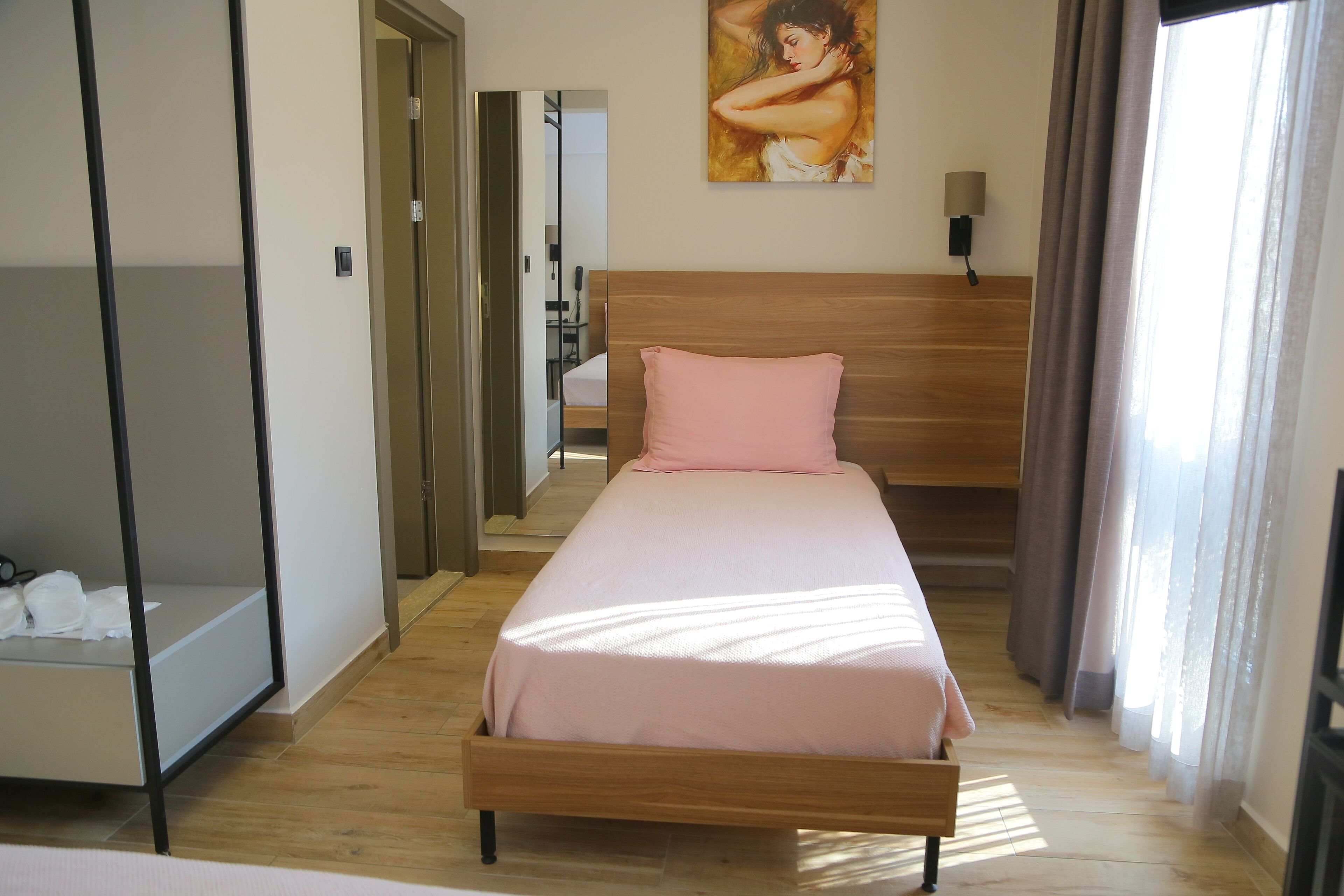 Une chambre d’hôtel avec un lit en bois, un oreiller rose, un tableau accroché au mur et une fenêtre avec des rideaux.