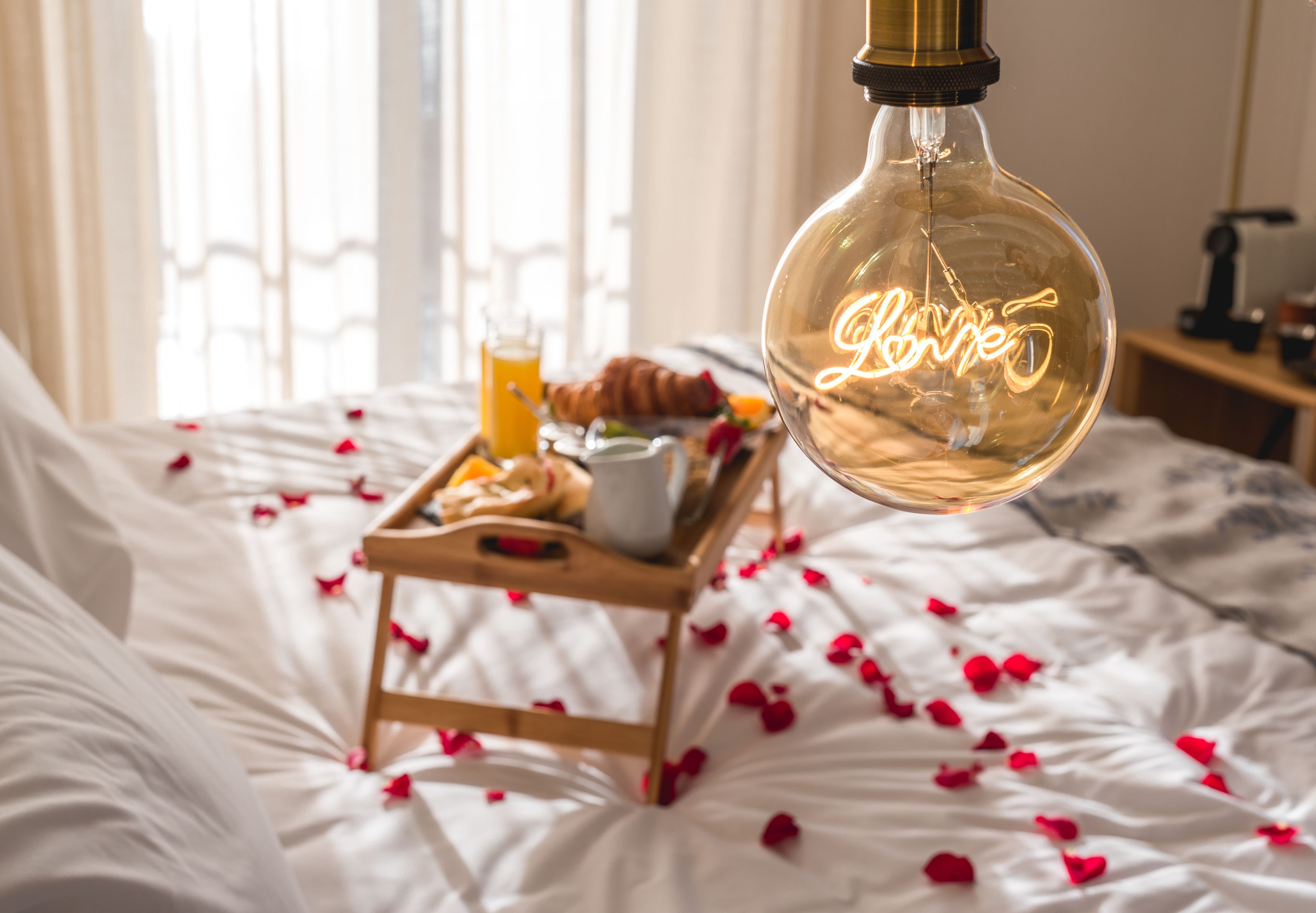A bed with rose petals, a tray with breakfast items, and a hanging light fixture with the word "love" in the room.