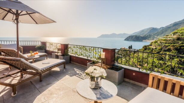 A terrace with lounge chairs, a table with flowers, and a view of the sea and hills.