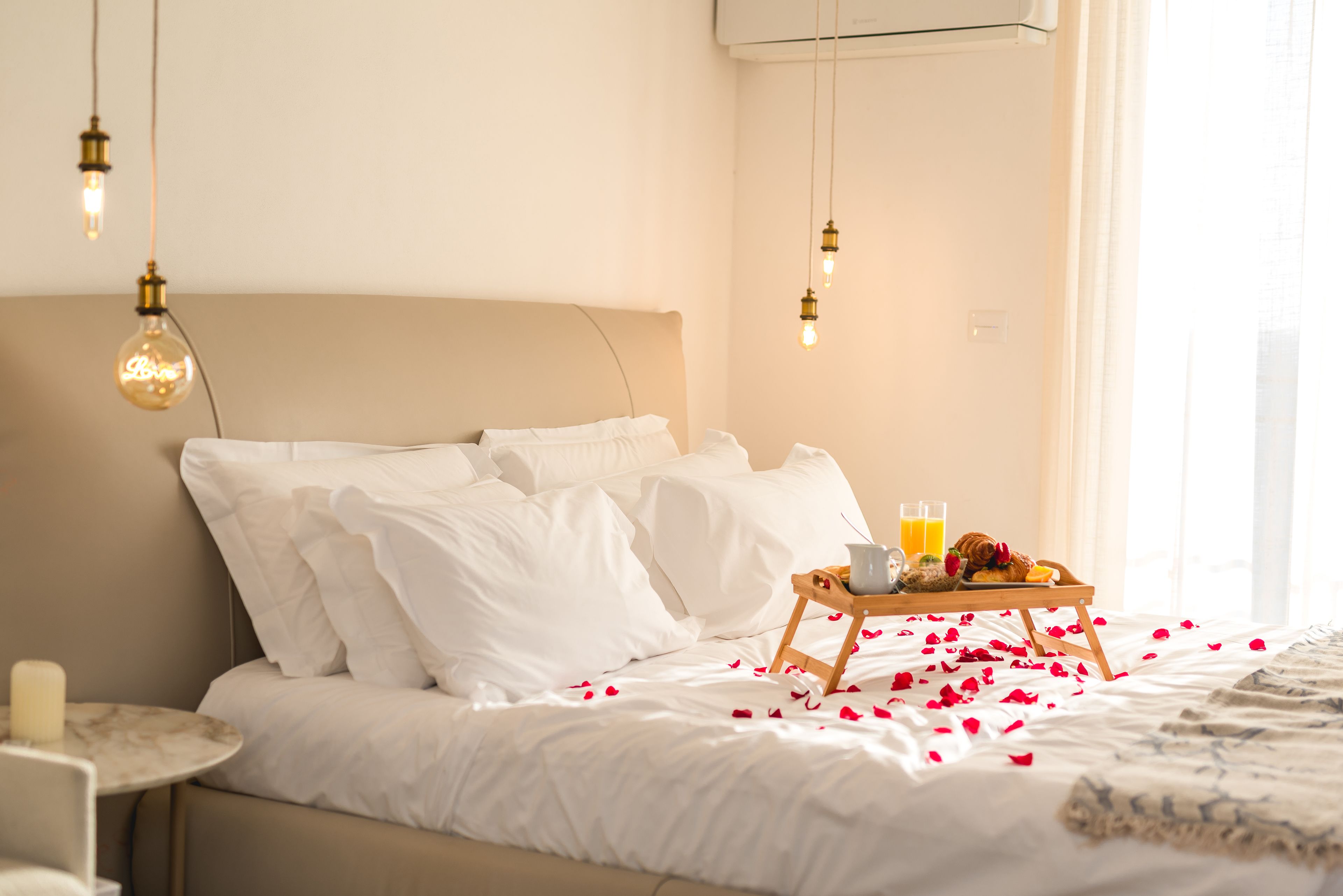 A neatly made bed with white linens, a tray of breakfast including toast and juice, and rose petals scattered on the bed.