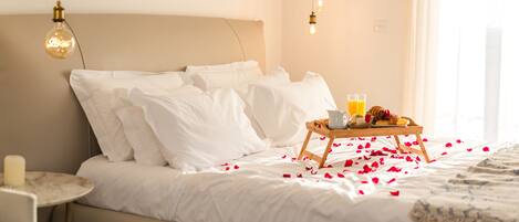 A neatly made bed with white linens, a tray of breakfast including toast and juice, and rose petals scattered on the bed.