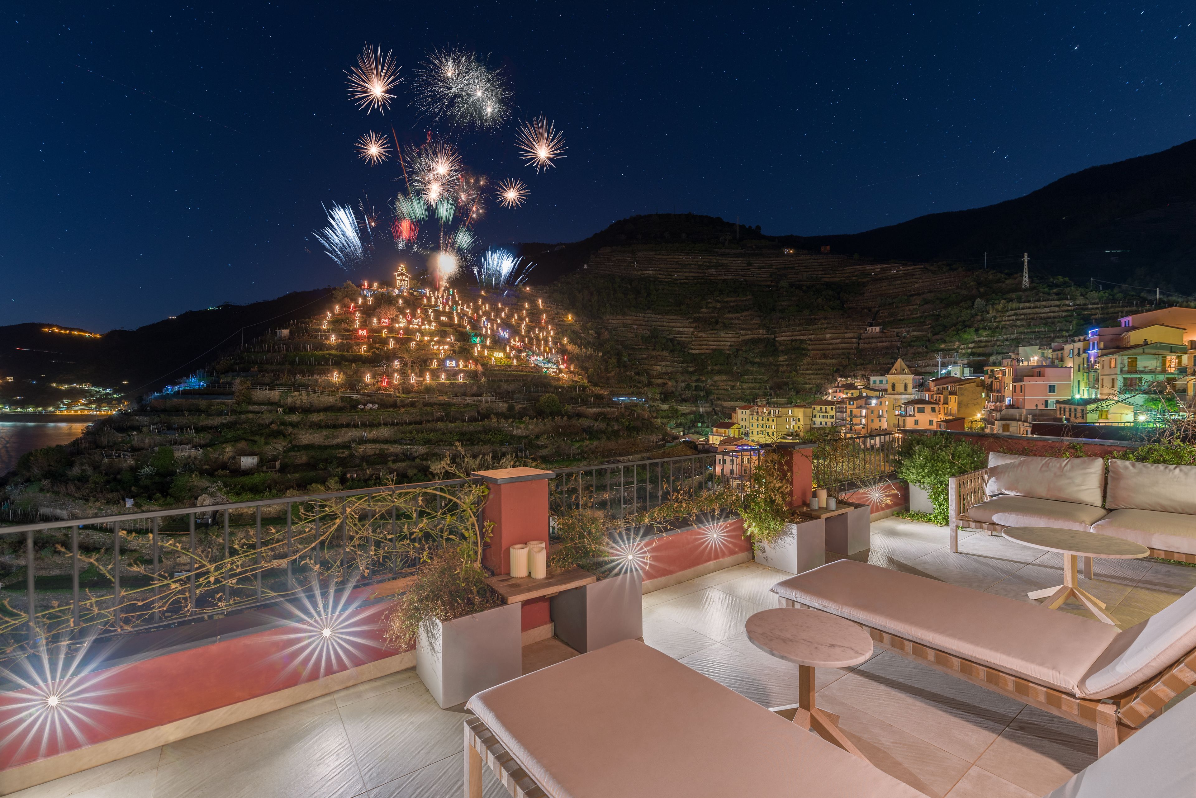 A rooftop terrace with lounge chairs and a view of a hillside village at night.