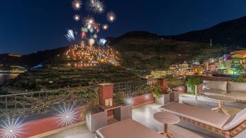 A rooftop terrace with lounge chairs and a view of a hillside village at night.