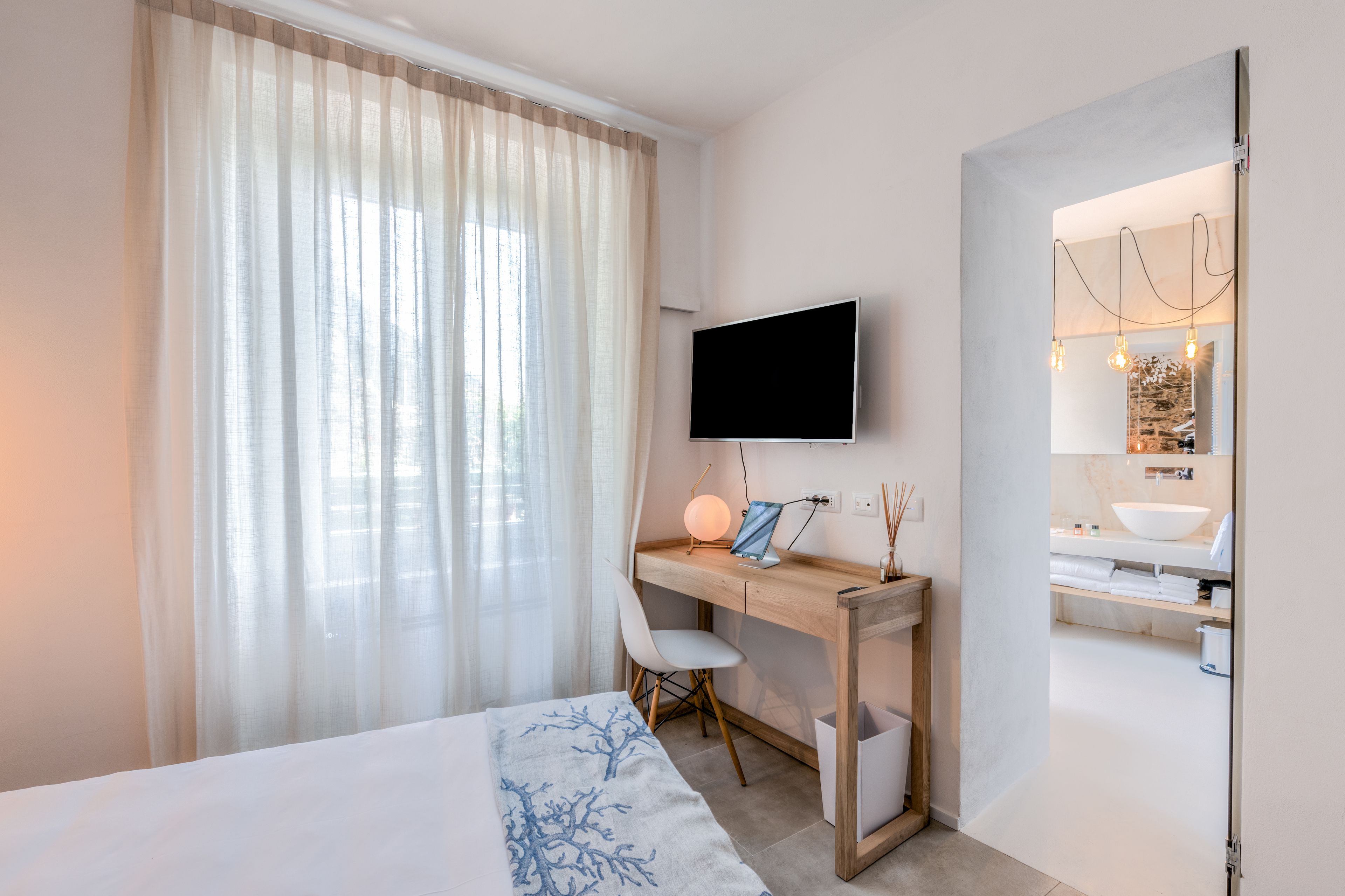 Camera d'albergo moderna con un letto, una scrivania con televisore, una sedia e un bagno visibile attraverso una porta aperta.