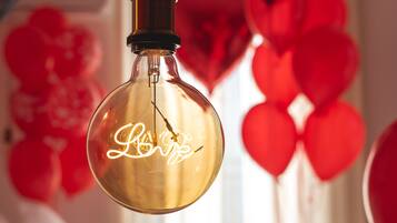 A round, clear glass light fixture with a visible filament, hanging in a room with red balloons and a heart-shaped decoration in the background.