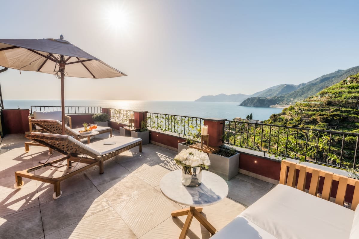 A sunlit terrace with lounge chairs, a table with flowers, and a view of the sea and hills.