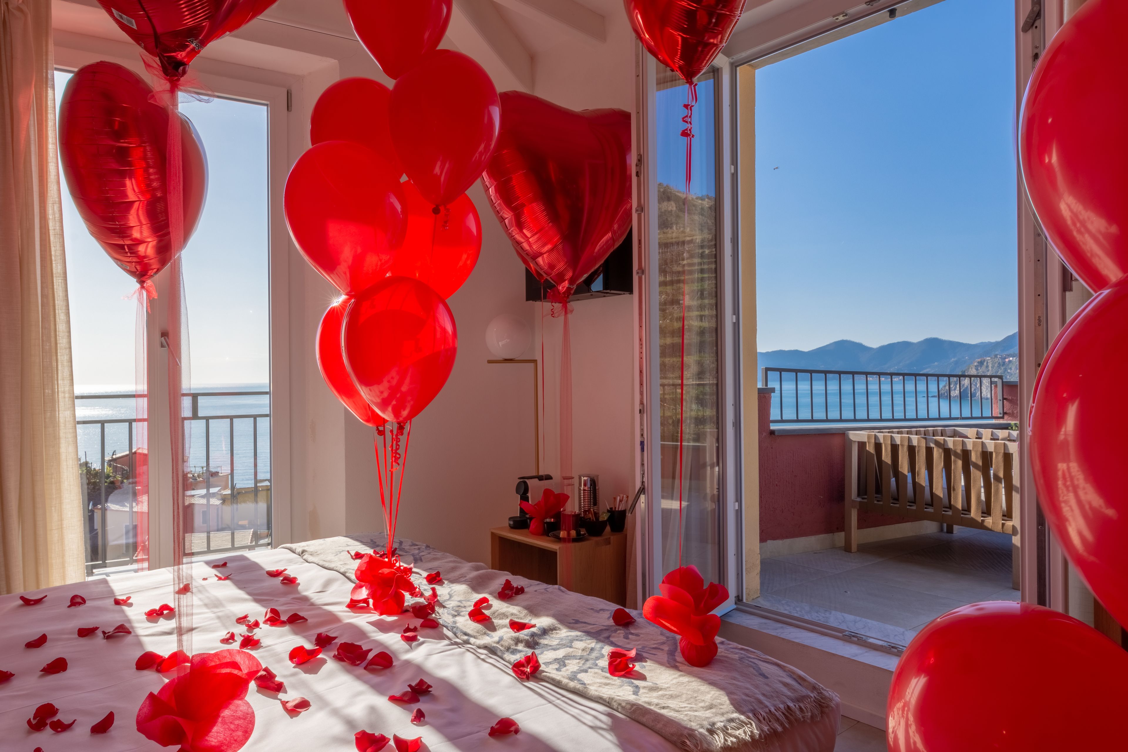 A room with a bed covered in rose petals, red heart-shaped balloons, and a balcony with a view of the sea and mountains.