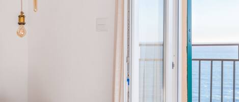Une chambre d’hôtel avec un lit, une table ronde et une vue sur la mer, accessible par une porte-fenêtre donnant sur un balcon.