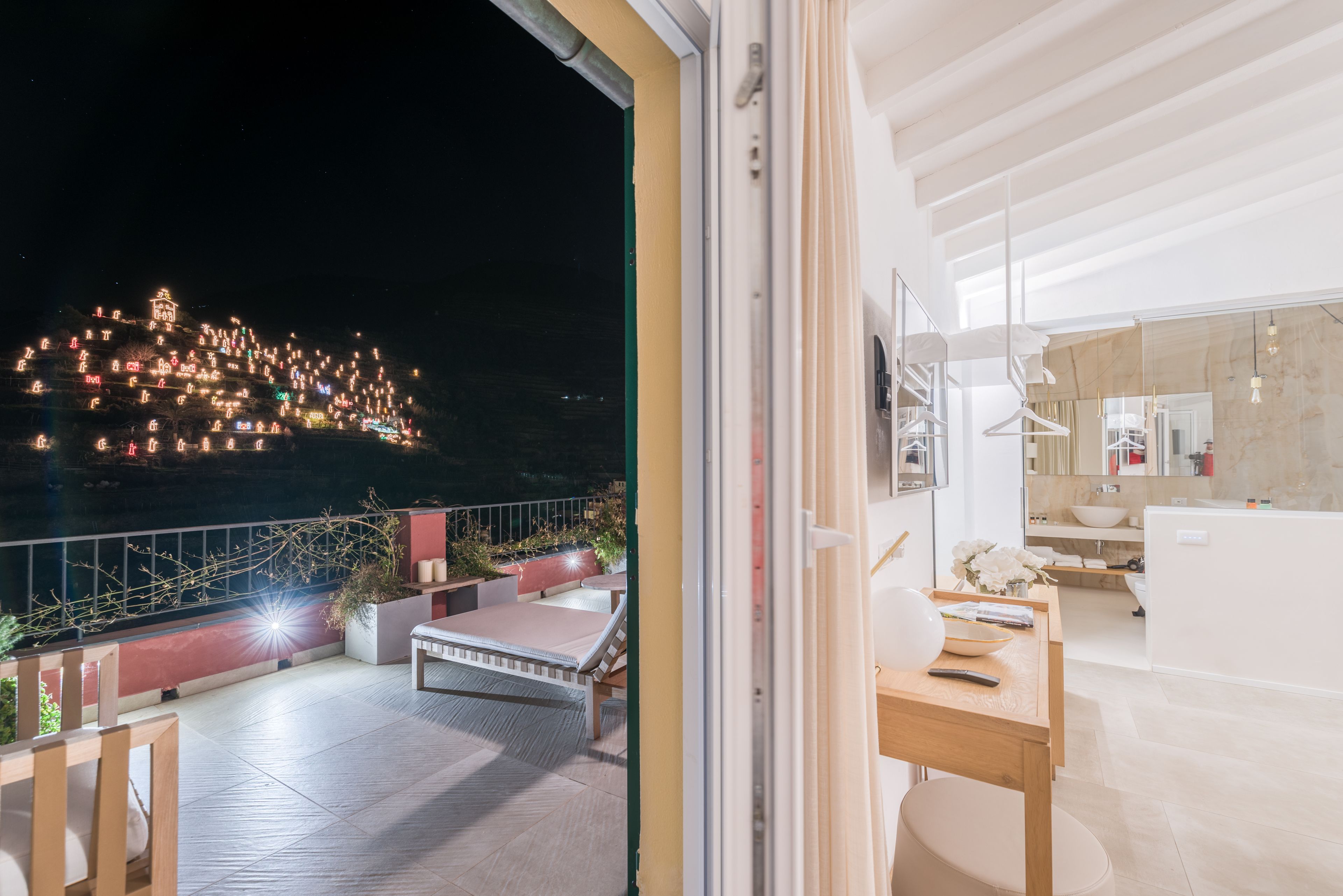 Un balcon offrant une vue sur une colline illuminée la nuit, un coin repas avec une table et des chaises, et une salle de bain moderne avec un grand miroir et un lavabo.
