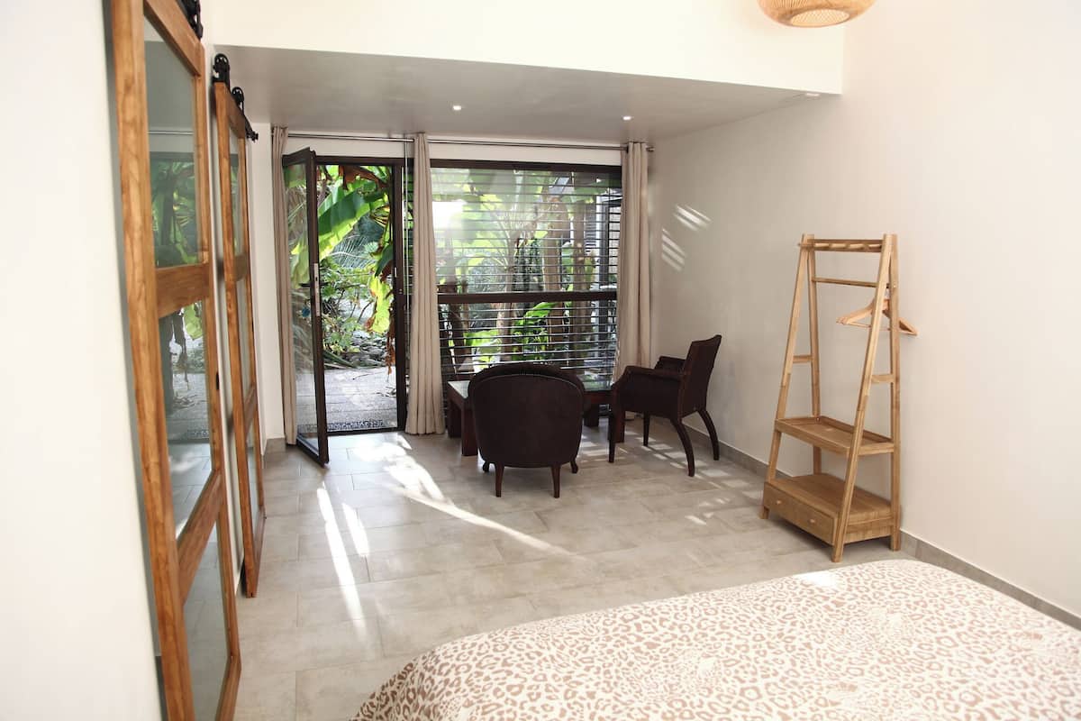 A room with a bed, a wooden chair, a small table, and a window with a view of greenery.