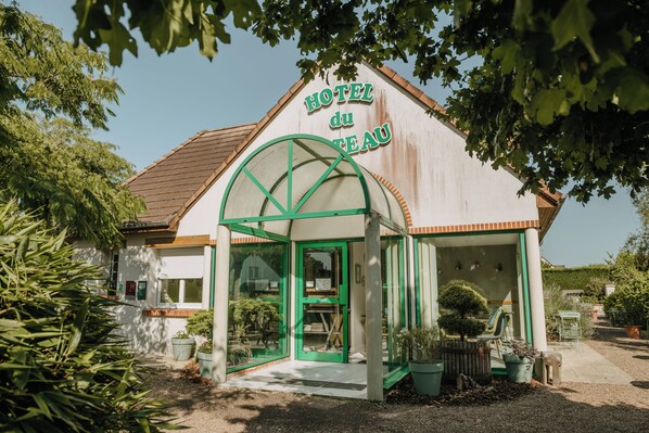 A quaint hotel with a distinctive green archway entrance, a sign reading "HOTEL du TEAU," and a small garden area with potted plants and a chair. - Hôtel du Chateau (Tour-en-Sologne)