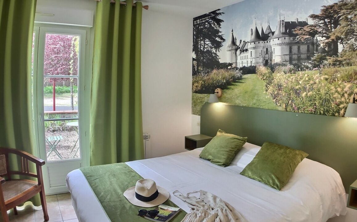 A hotel room with a bed, green curtains, a window, a chair, and a large painting of a castle.