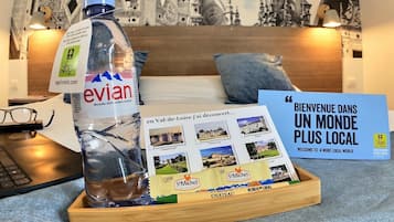 Habitación de hotel con cama, ordenador portátil, botella de agua, folleto y bandeja de madera con folleto y tableta de chocolate.