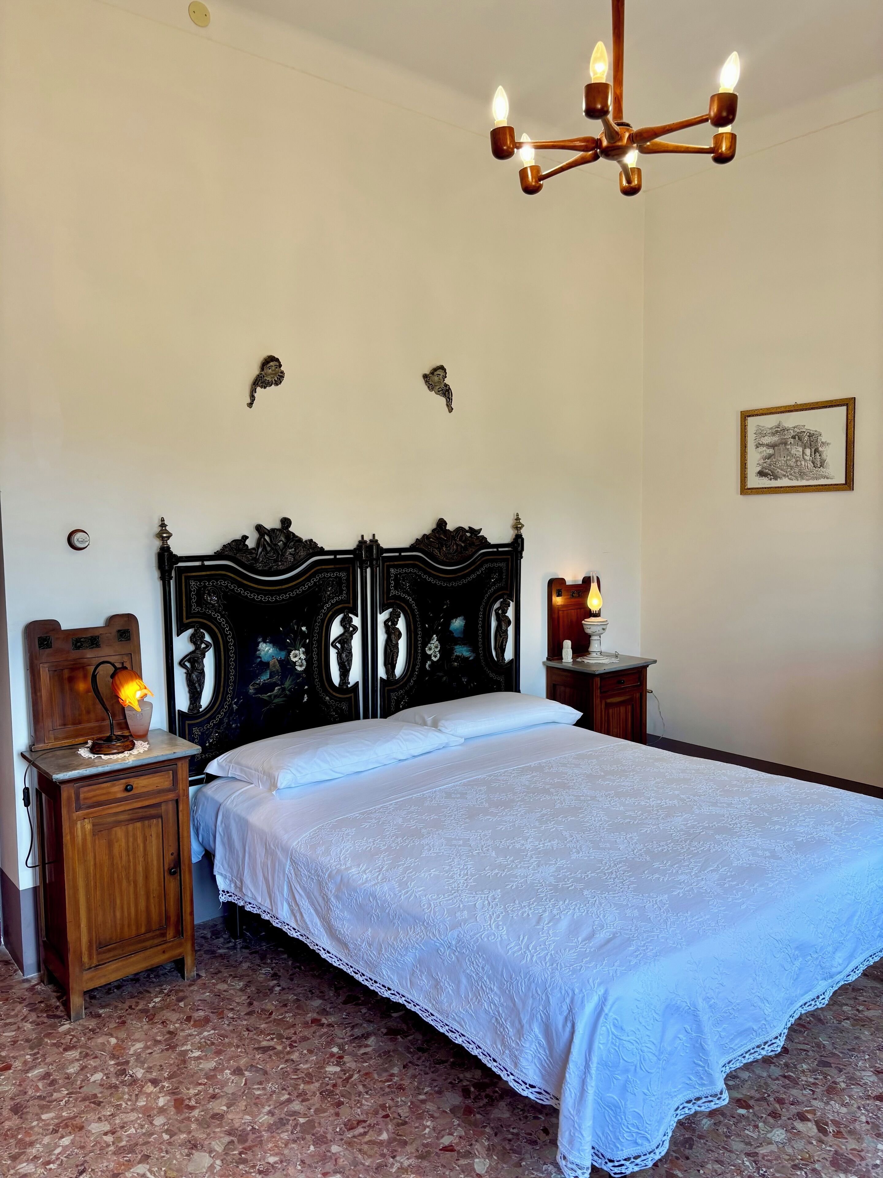 A bedroom with a large bed, a wooden nightstand with a lamp, a wall-mounted chandelier, and a framed picture on the wall.