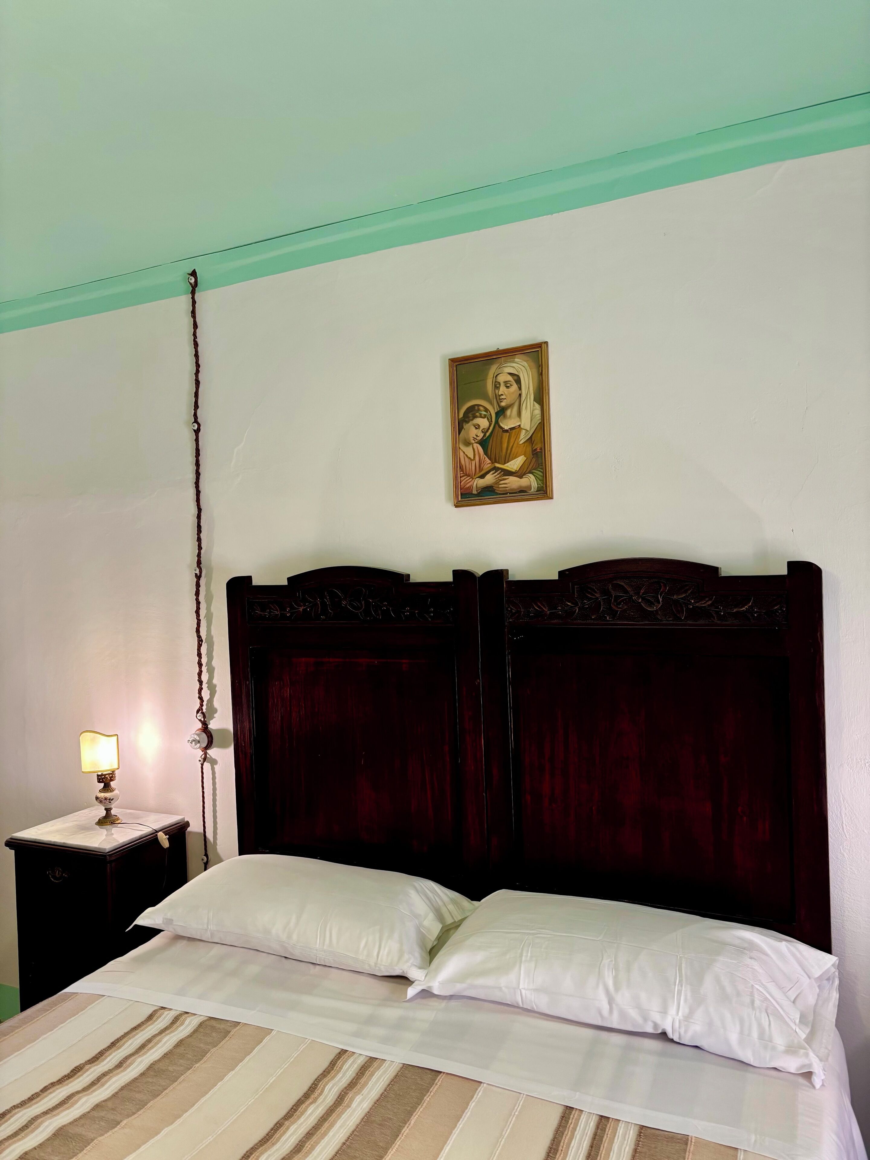 Quarto com cabeceira de madeira, cama com roupa de cama branca, mesa de cabeceira com abajur e um objeto decorativo na parede.