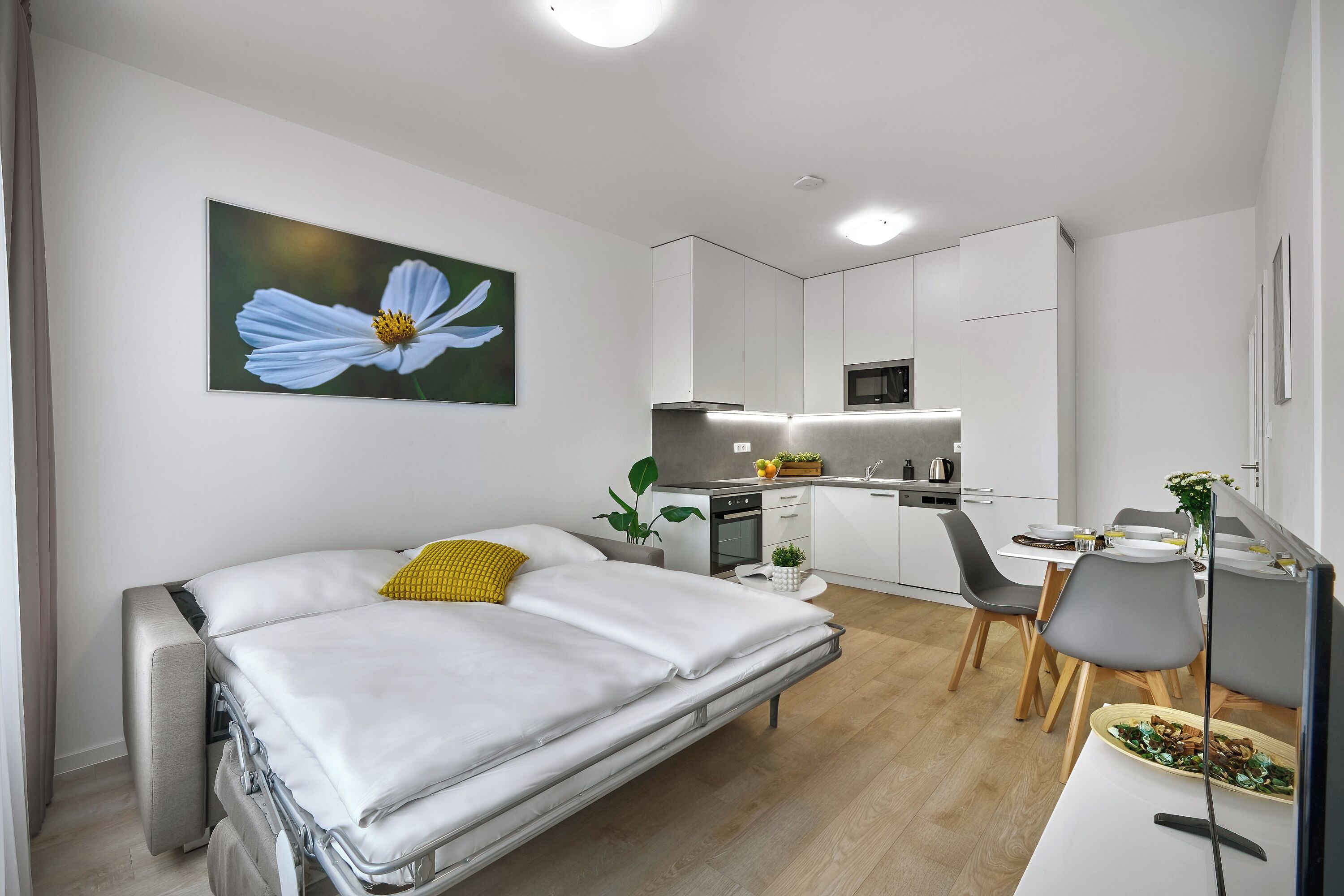 Une chambre moderne avec un lit, un coin repas avec des chaises, une cuisine avec des placards et un grand tableau représentant des fleurs accroché au mur.