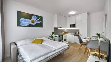 Une chambre moderne avec un lit, un coin repas avec des chaises, une cuisine avec des placards et un grand tableau représentant des fleurs accroché au mur.