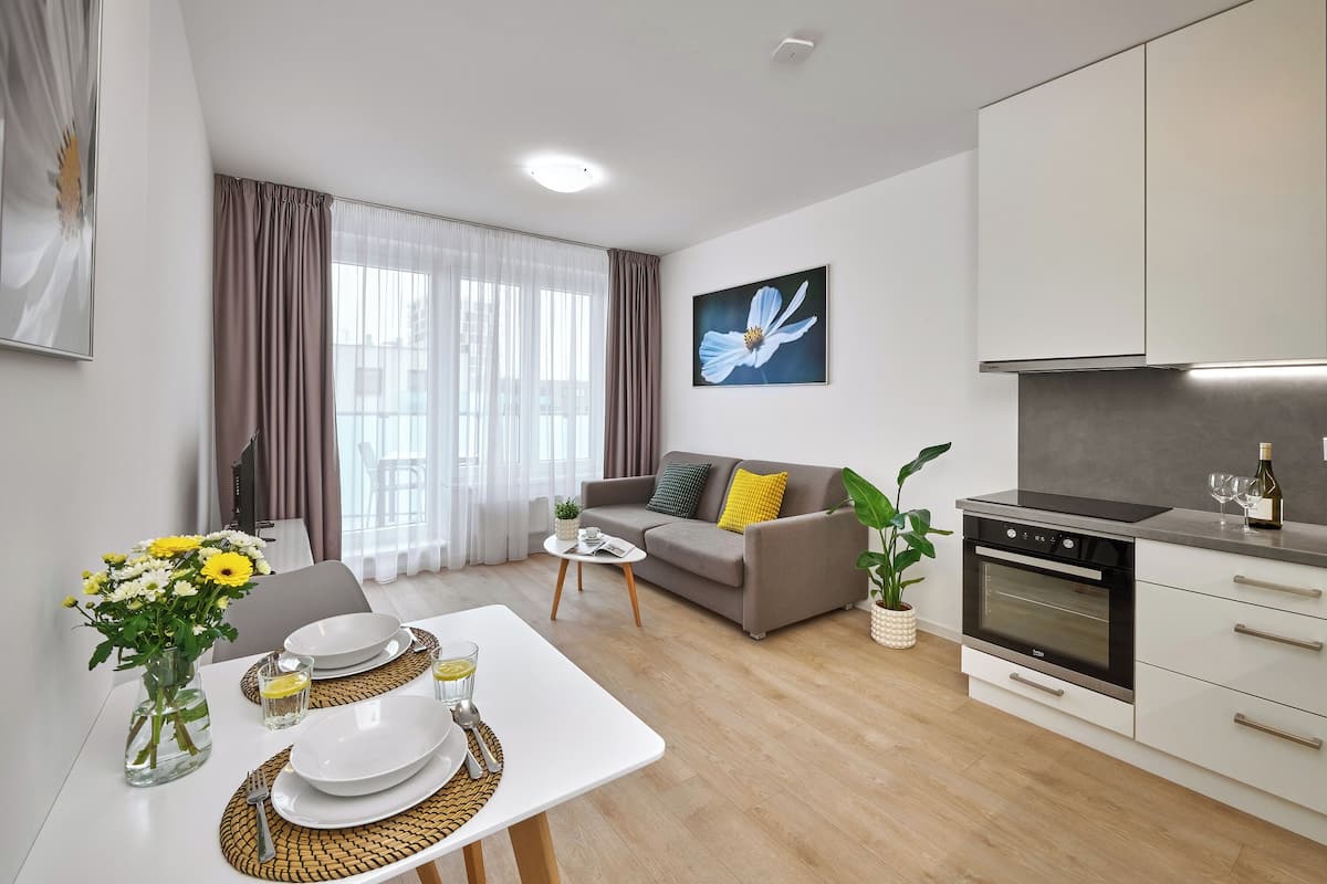 A modern living room with a dining table set for two, a sofa with cushions, a kitchen with a stove, and a large window with curtains.
