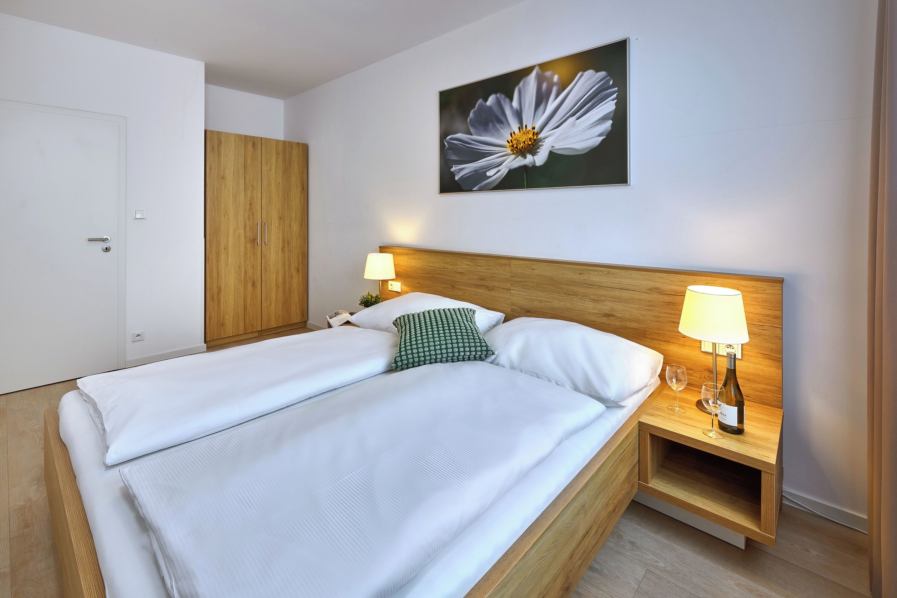 A neatly made bed with white linens, a wooden headboard, and a nightstand with a lamp and a bottle.