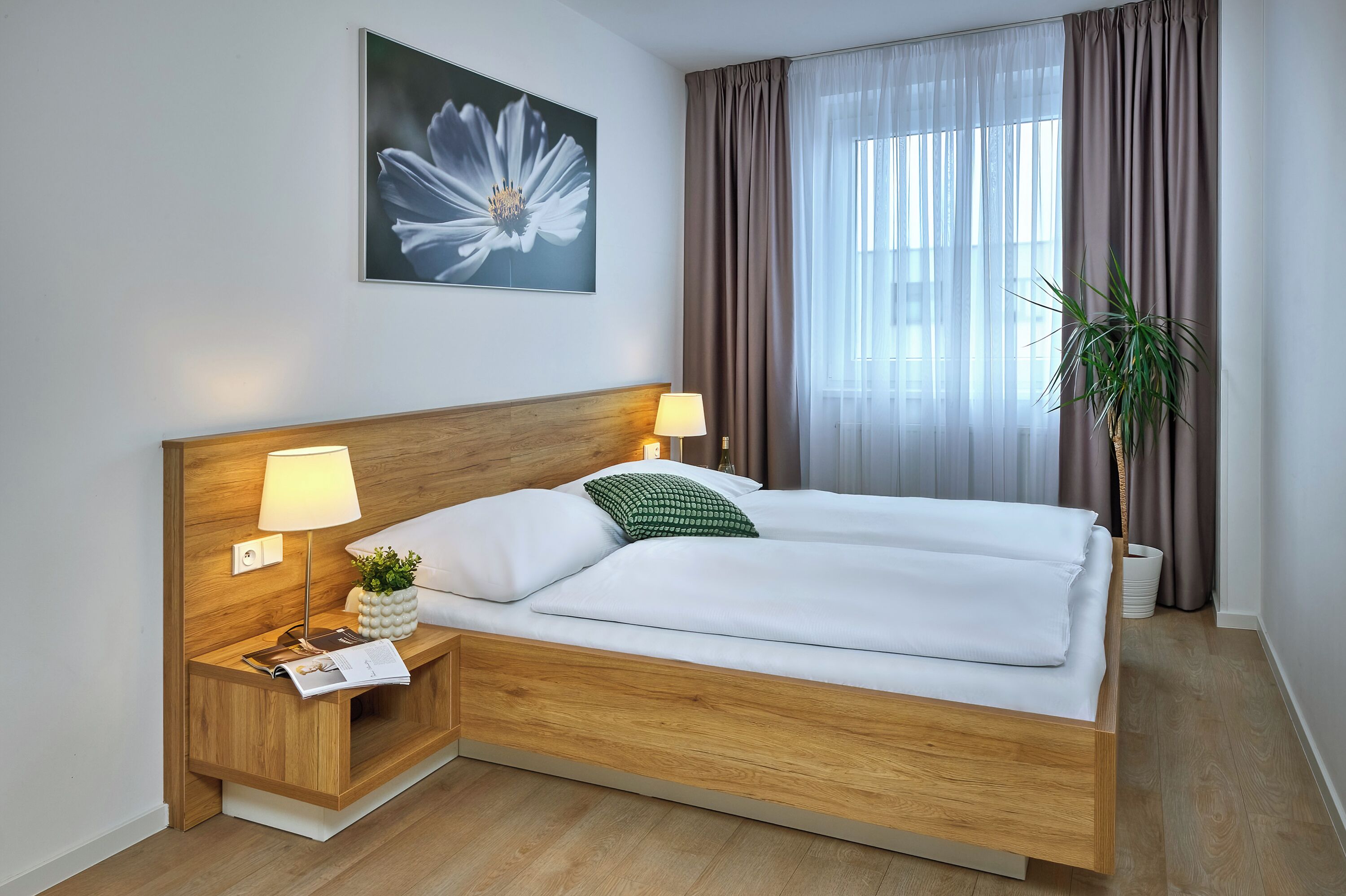 A neatly made bed with white linens, two bedside lamps, a wooden nightstand with a plant, and a large window with curtains.