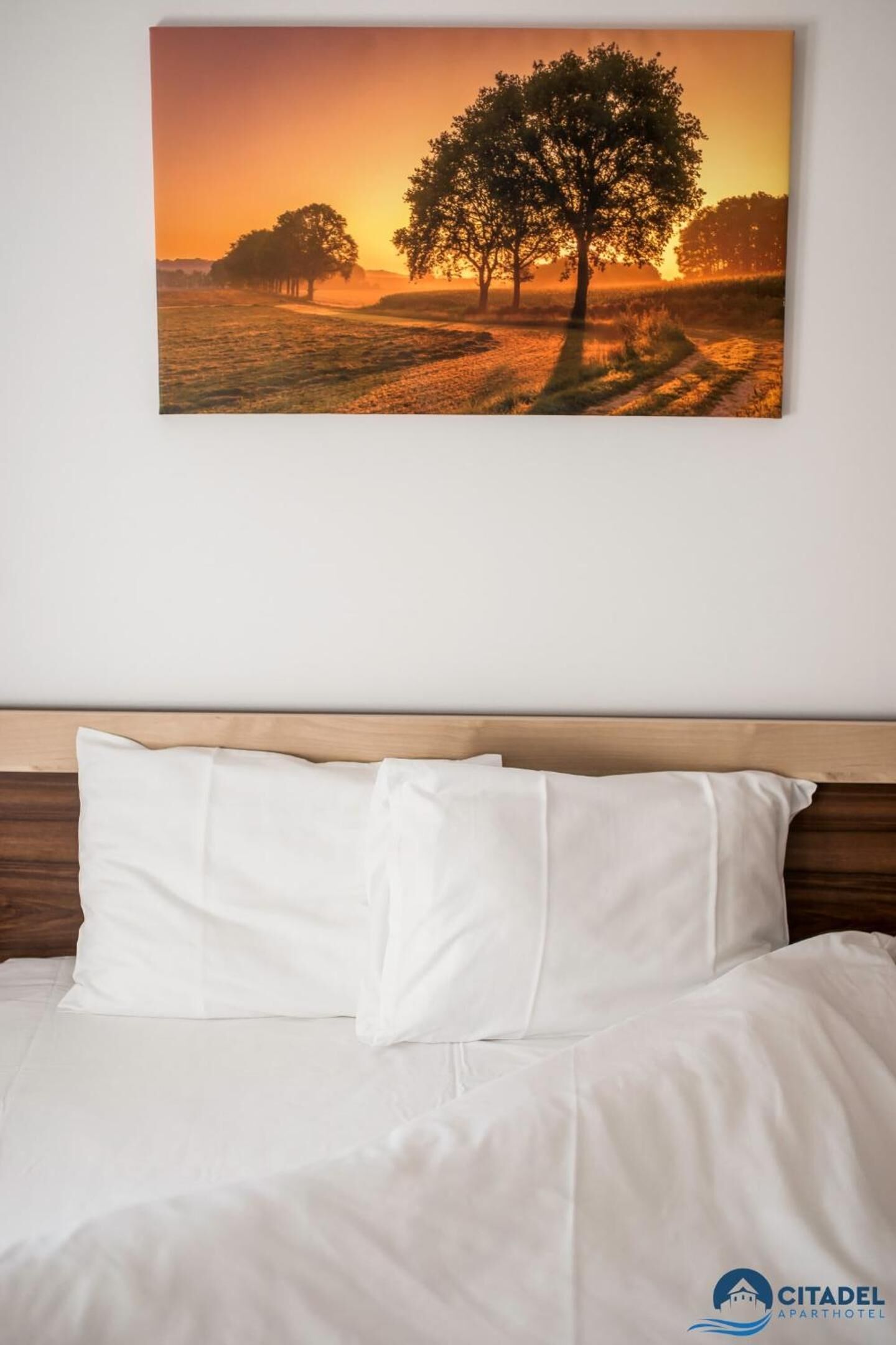 A bed with white bedding and pillows, a wooden headboard, and a landscape painting of a sunset with trees on the wall.