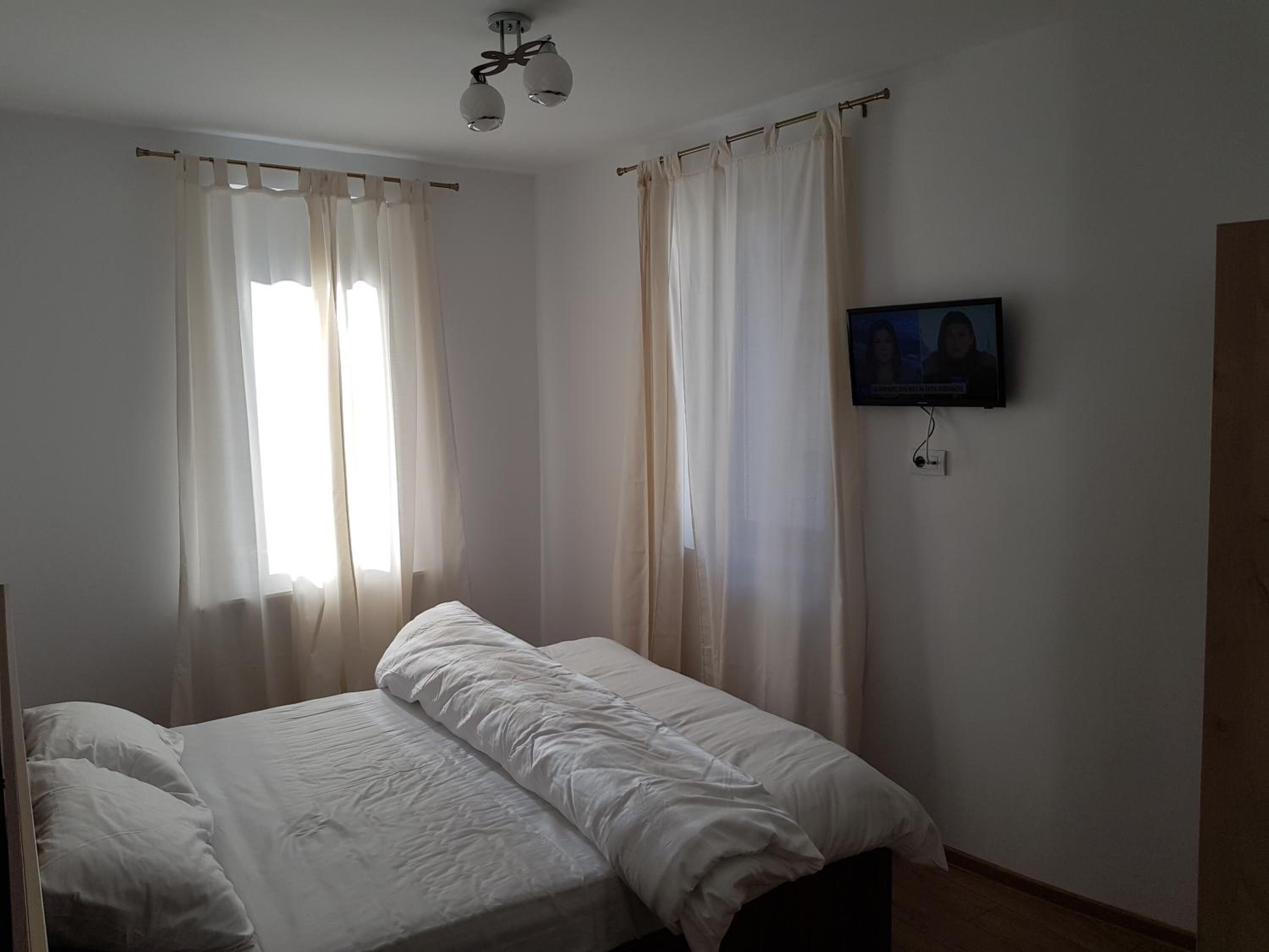 Een slaapkamer met een bed, gordijnen, een plafondlamp en een televisie aan de muur.
