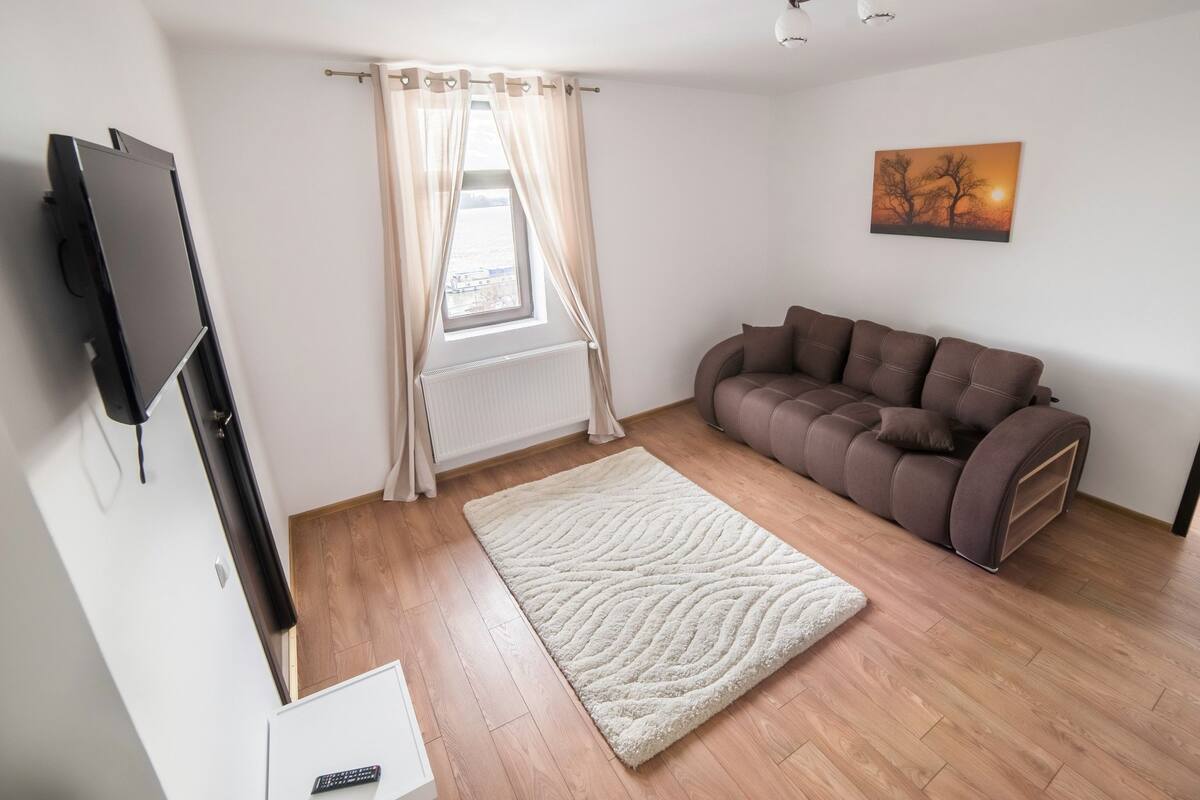 A living room with a brown sofa, a white rug, a TV mounted on the wall, and a window with curtains.
