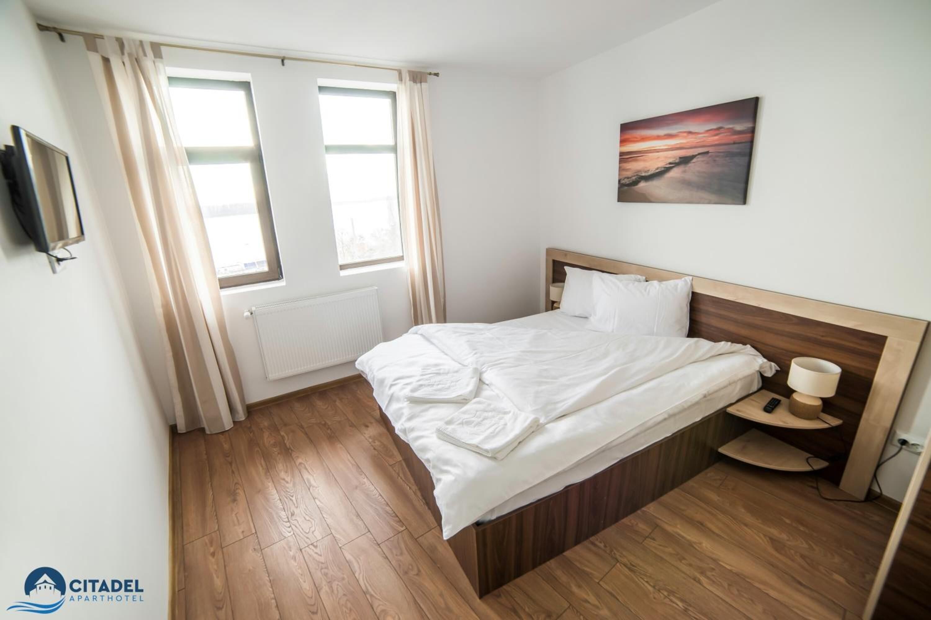 Hotellihuone, jossa on puulattia, valkoisilla lakanoiden peitetty sänky, yöpöytä lampun kanssa ja taulu seinällä.