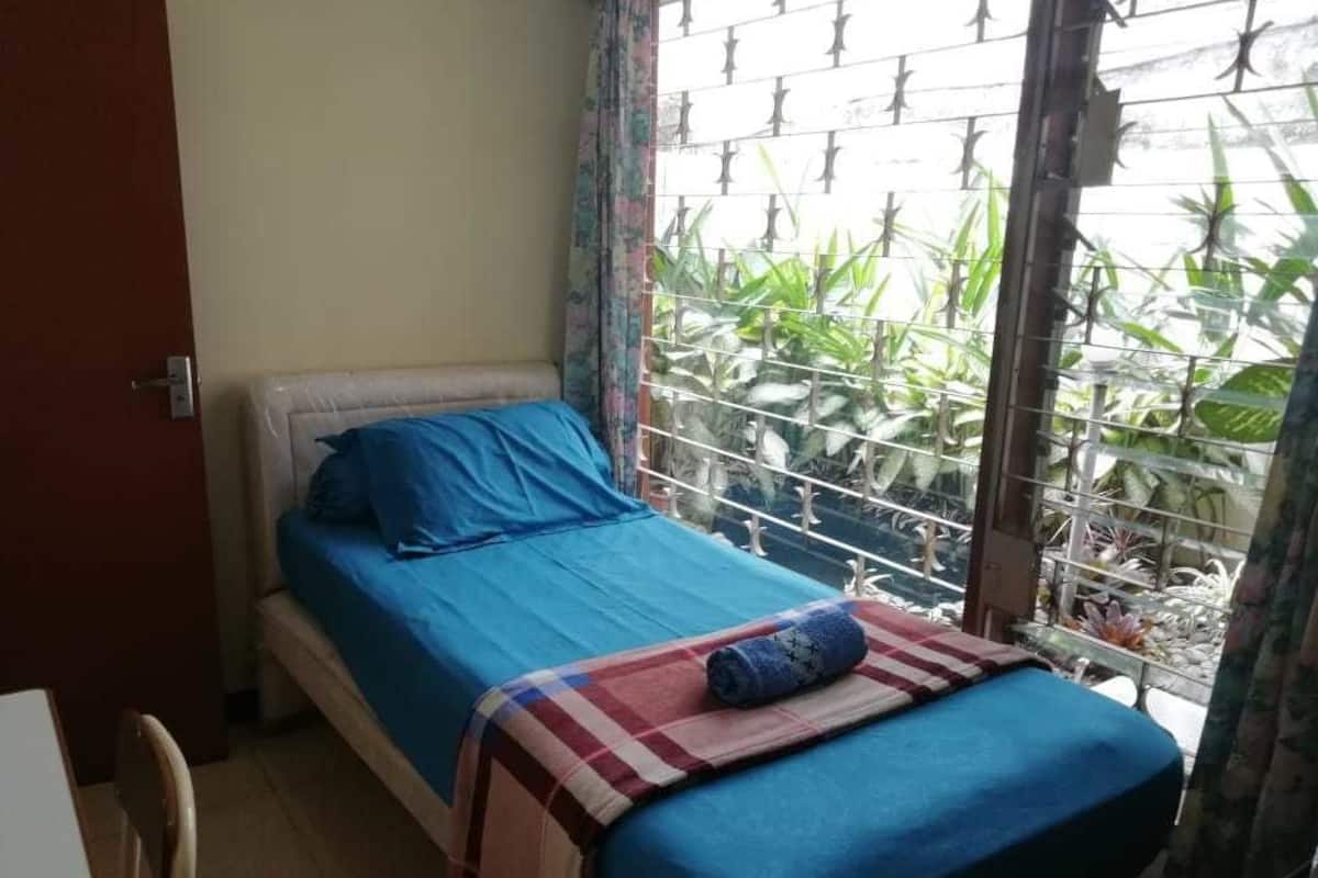 A single bed with blue bedding, a red and white checkered blanket, and a rolled towel. A window with metal bars and green plants is visible.