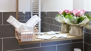 Una encimera de baño con una cesta metálica que contiene bolsas blancas y papeles, y un florero con flores rosas.