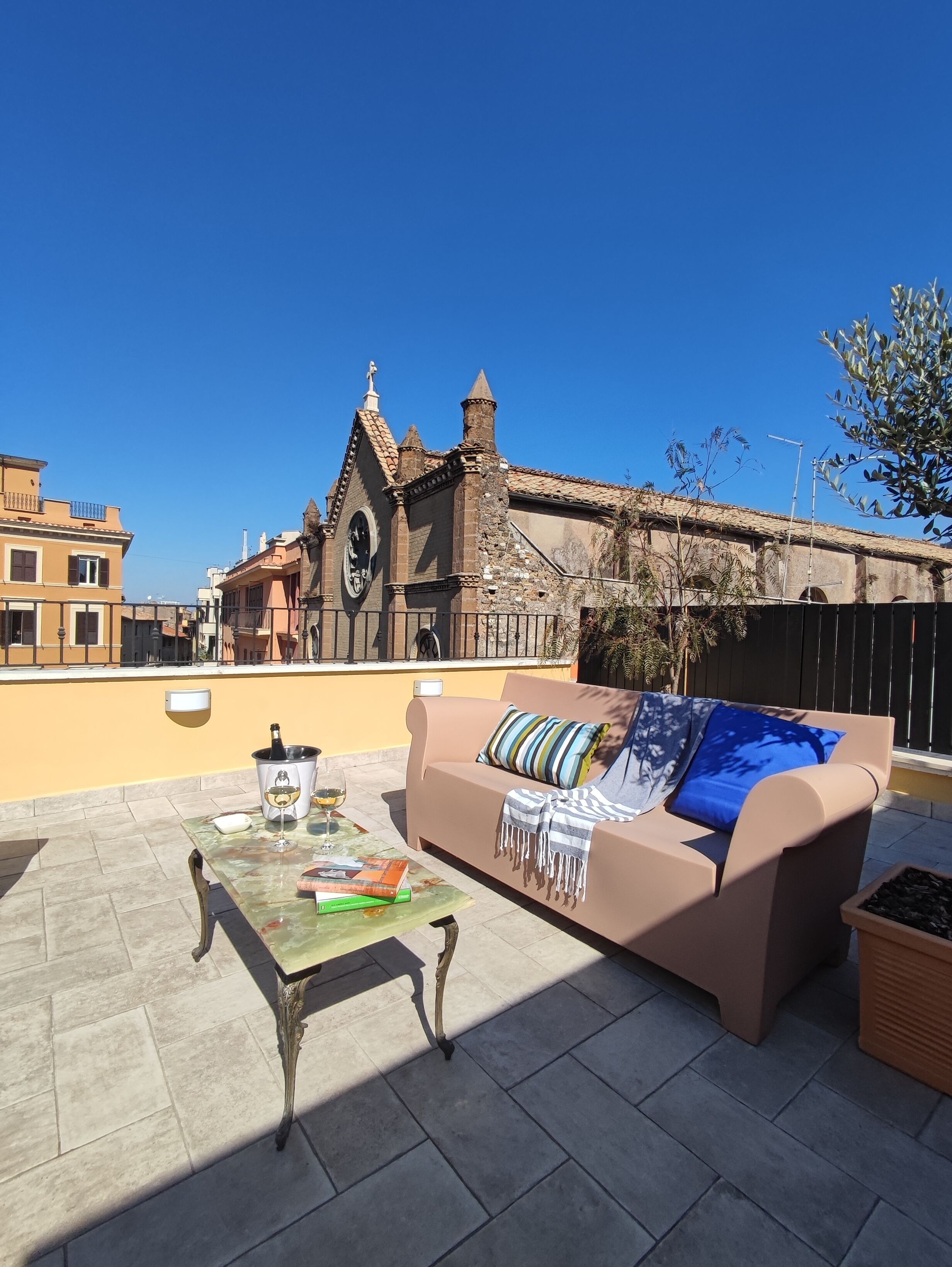 A rooftop terrace with a beige sofa, a table with a bottle of champagne, and a book. In the background, there is a building with a clock tower and a church.