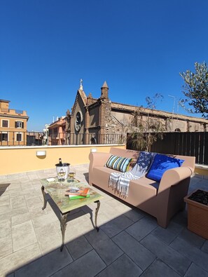 A rooftop terrace with a beige sofa, a table with a bottle of champagne, and a book. In the background, there is a building with a clock tower and a church.