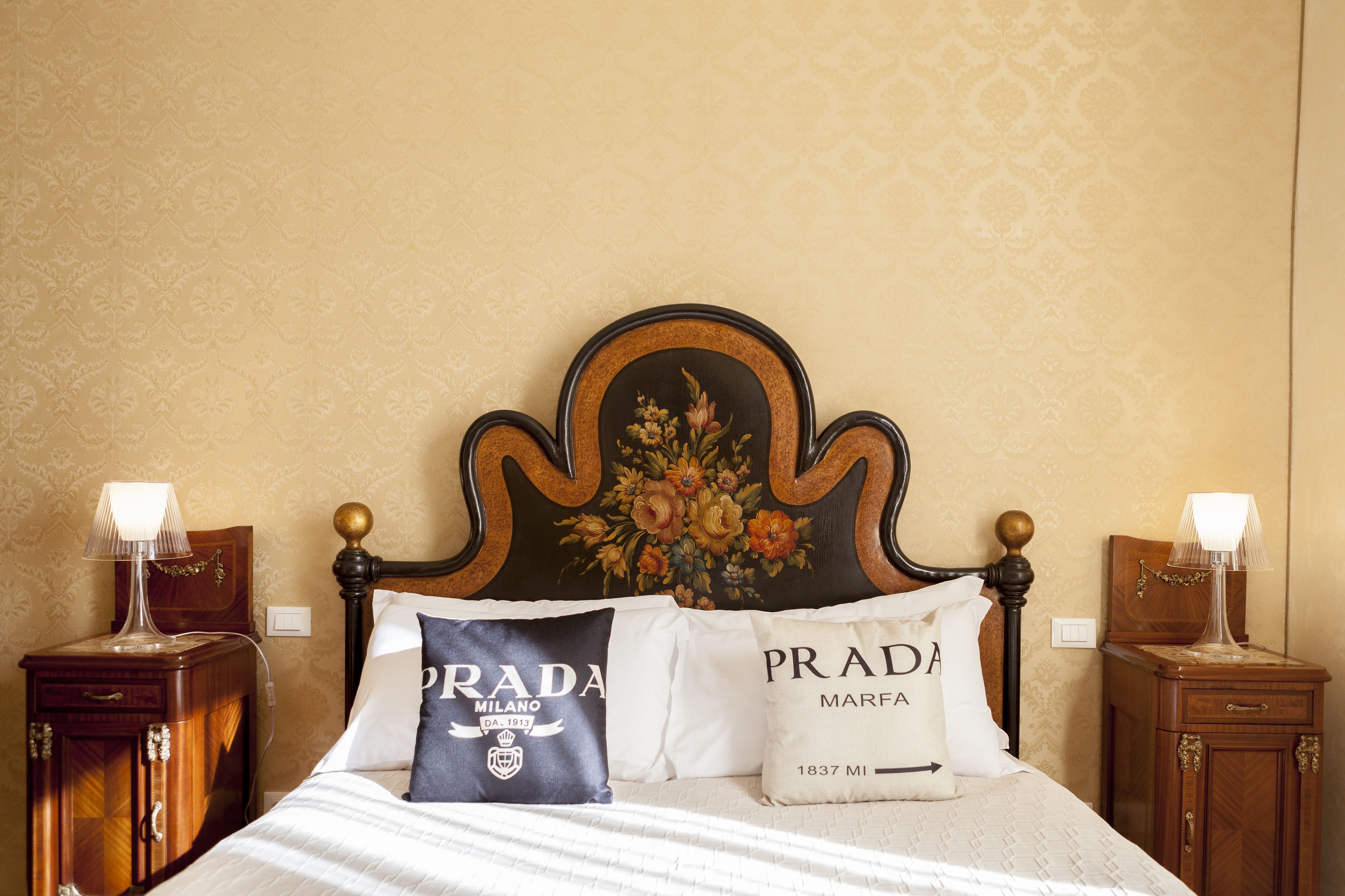 A bed with a headboard featuring a floral design, two pillows with "PRADA MILANO" and "PRADA MARFA" text, and two bedside tables with lamps.