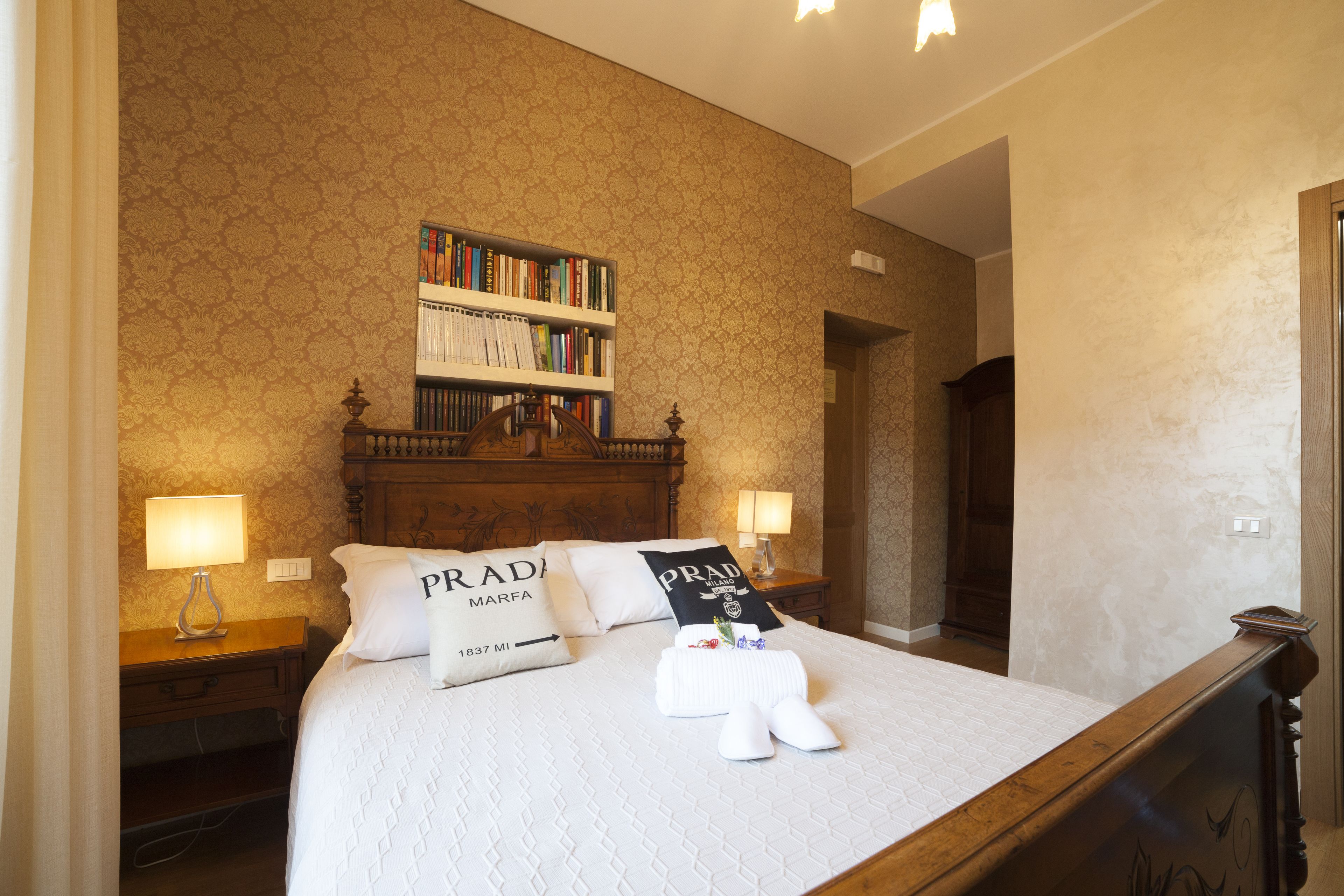 A neatly made bed with a white bedspread and two pillows, one with "PRADA MARFA" and another with "PRADA" text. There is a bookshelf above the bed with various books, a wooden headboard, and a nightstand with a lamp.
