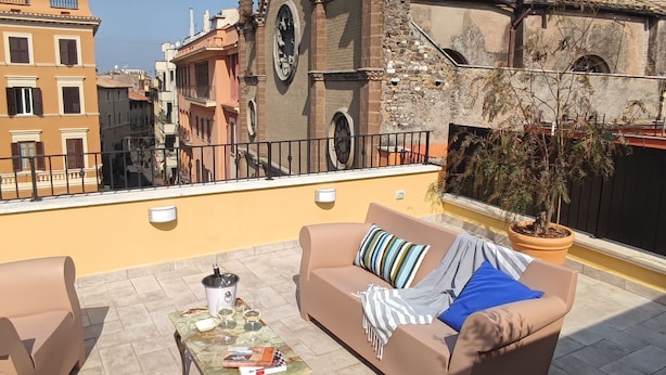 Una terraza en la azotea con sofá y sillones, una mesa con bebidas y libros, y vistas a edificios históricos.